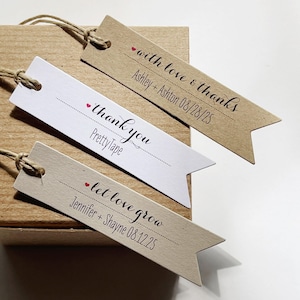 May include: Three brown and white paper gift tags with a red heart and black text. The text on the tags reads "with love & thanks Ashley + Ashton 08/28/25", "thank you Pretty Tape", and "let love grow Jennifer + Shayne 08.12.25".