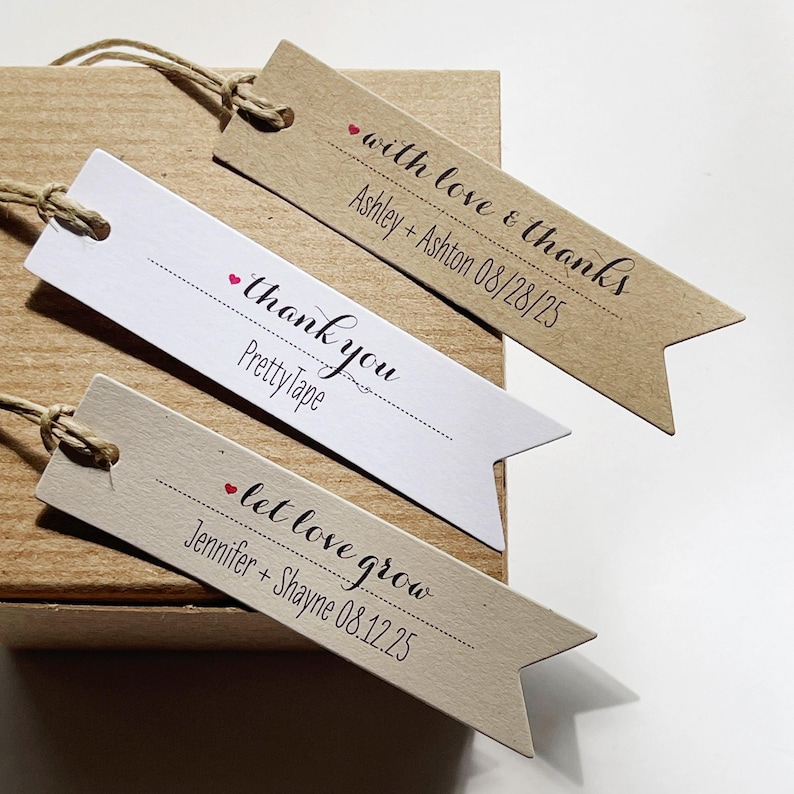 May include: Three brown and white paper gift tags with a red heart and black text. The text on the tags reads "with love & thanks Ashley + Ashton 08/28/25", "thank you Pretty Tape", and "let love grow Jennifer + Shayne 08.12.25".