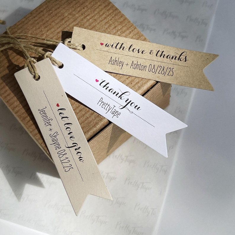May include: Three brown and white paper gift tags with a red heart and black text. The text on the tags reads "with love & thanks Ashley + Ashton 08/28/25", "thank you Pretty Tape", and "let love grow Jennifer + Shayne 08.12.25".