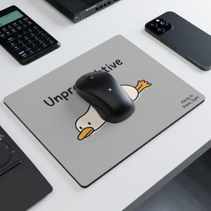 May include: A grey mouse mat featuring the word "Unproductive" and a cartoon duck design. A black wireless mouse rests on the mat. The text "Hang in there, Sam" is also present. Other electronics are in the background.