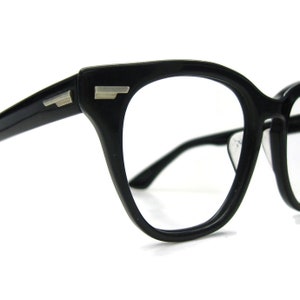 Puede incluir: Gafas de plástico negro con detalles plateados en las patillas. Las gafas tienen forma de ojo de gato.