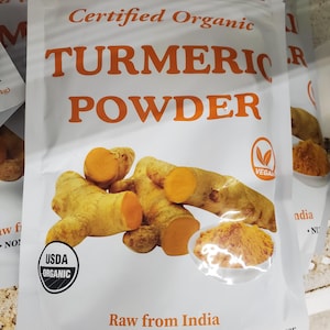May include: A white bag of turmeric powder with the words "TURMERIC POWDER" in orange. The bag also features images of turmeric root and powder, and states "USDA ORGANIC", "GLUTEN FREE", and "NON-GMO". The net weight is 16 oz (454g).