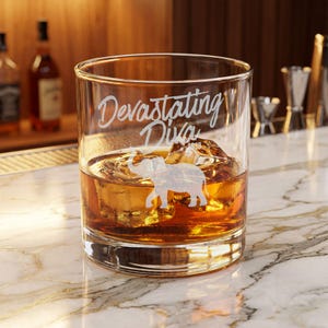 May include: A clear glass tumbler filled with amber liquid and ice cubes. The glass features an etched design of an elephant and the text "Devastating Diva" in a script font. The glass has a thick base and sits on a white marble surface.