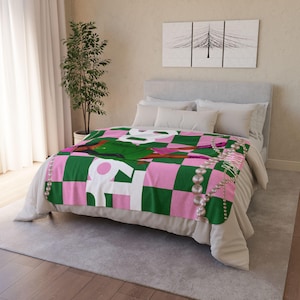 Op de afbeelding: Een groen-roze geruite deken met een parelkettingontwerp en een afbeelding van een vrouw in een groene jurk. De deken ligt op een bed met witte en grijze kussens.