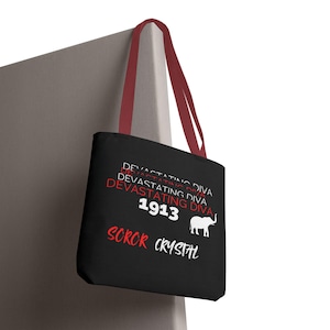 May include: Black tote bag with burgundy handles. The bag features the text "DEVASTATING DIVA" in red and white, along with the year "1913" and a white elephant graphic. The text "SOROR CRYSTAL" is also displayed.