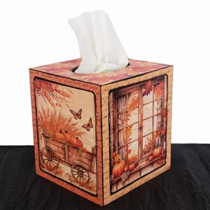 May include: A wooden tissue box cover with a fall theme. The design features a window with fall leaves, a wagon full of pumpkins, and butterflies. The box is painted in shades of orange, brown, and yellow.