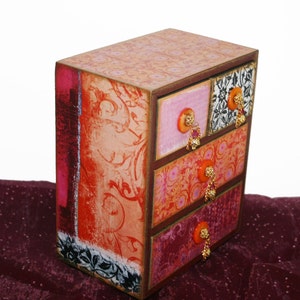 May include: A small wooden box with four drawers, each decorated with a different patterned paper. The drawers have small, decorative knobs with dangling beads.