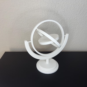May include: A white, three-dimensional sculpture with a spherical design. The sculpture features a circular frame with intersecting rings and a central disc. The base is a circular pedestal. The object is set against a neutral background.