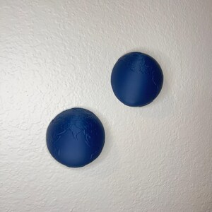 K&ouml;nnte beinhalten: Zwei marineblaue, globusf&ouml;rmige Wanddekorationen. Die Globen haben eine strukturierte Oberfl&auml;che mit erhabenen Kontinenten und sind an einer wei&szlig;en Wand befestigt. Die Globen haben einen Durchmesser von etwa 10 cm.