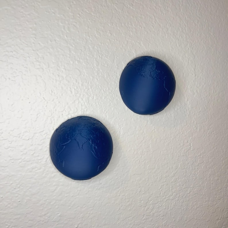 K&ouml;nnte beinhalten: Zwei marineblaue, globusf&ouml;rmige Wanddekorationen. Die Globen haben eine strukturierte Oberfl&auml;che mit erhabenen Kontinenten und sind an einer wei&szlig;en Wand befestigt. Die Globen haben einen Durchmesser von etwa 10 cm.