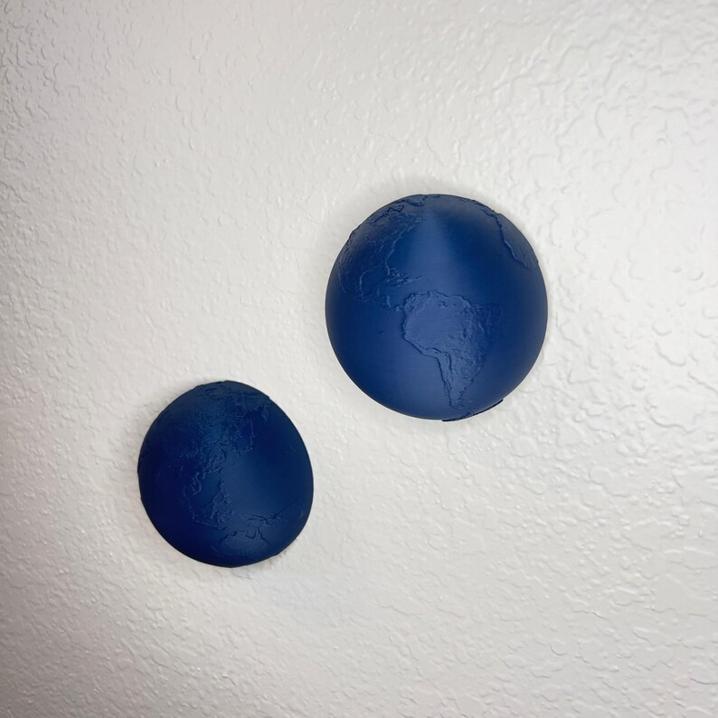 K&ouml;nnte beinhalten: Zwei marineblaue, globusf&ouml;rmige Wanddekorationen. Der gr&ouml;&szlig;ere Globus zeigt Kontinente und Ozeane im Relief. Der kleinere Globus ist ebenfalls marineblau und hat eine &auml;hnliche strukturierte Oberfl&auml;che. Beide sind an einer strukturierten wei&szlig;en Wand befestigt.