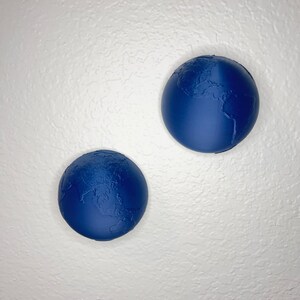 K&ouml;nnte beinhalten: Zwei blaue Globen mit detaillierten Landmassenmerkmalen sind an einer strukturierten wei&szlig;en Wand befestigt. Die Globen sind in einem tiefen Blauton gehalten, und die Kontinente sind klar definiert, wodurch ein dreidimensionaler Effekt entsteht.