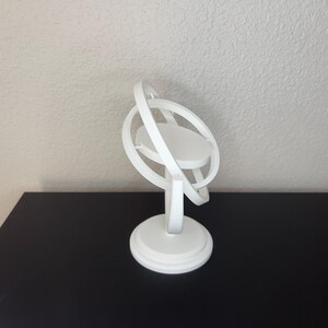 May include: A white, 3D-printed armillary sphere model. The model features a circular base, a central platform, and multiple orbiting rings. The design is minimalist and modern, suitable for display or educational purposes.