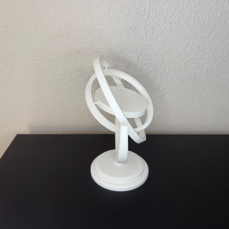 May include: A white, 3D-printed armillary sphere model. The model features a circular base, a central platform, and multiple orbiting rings. The design is minimalist and modern, suitable for display or educational purposes.