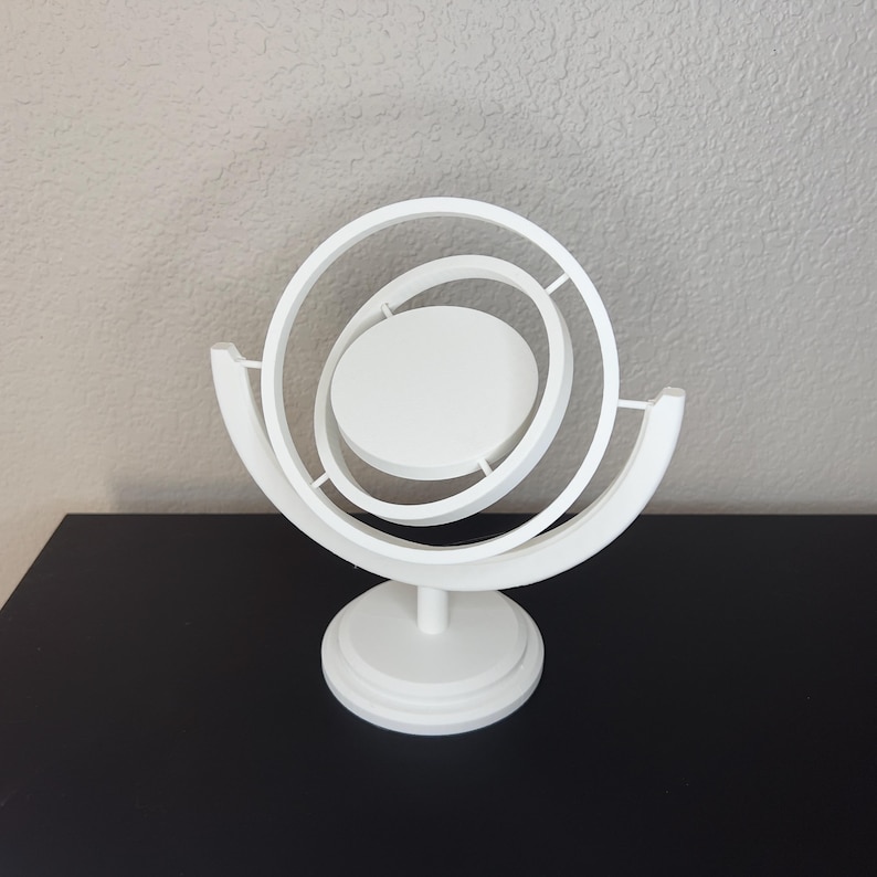 May include: A white, abstract sculpture with a circular design. The sculpture features concentric rings and a central disc, all supported by a pedestal base. The piece is made of a solid, opaque material and sits on a black surface.