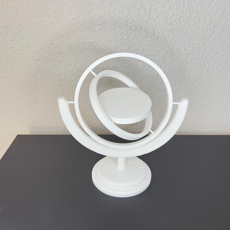 May include: A white, 3D-printed decorative sculpture featuring a central circular platform surrounded by orbiting rings. The sculpture is supported by a curved base and a circular pedestal, creating a modern, geometric design. The piece is displayed on a dark surface.