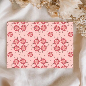 Könnte beinhalten: Eine rechteckige Karte mit einem sich wiederholenden Muster aus rosa und roten floralen Designs auf einem hellrosa Hintergrund. Das florale Muster umfasst kreisförmige und sternförmige Formen mit kleinen blattartigen Details. Die Karte liegt auf einem weißen Stoff.