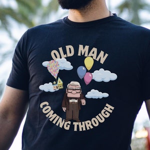 Puede incluir: Camiseta negra con el texto "OLD MAN COMING THROUGH" y un dibujo animado de una casa, globos y un anciano. La camiseta la lleva una persona con barba.