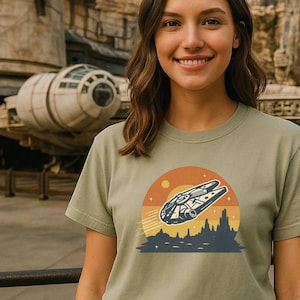Può includere: Una donna indossa una maglietta verde con una grafica della navicella spaziale Millennium Falcon contro un tramonto. Indossa anche orecchie da topo nere. Lo sfondo presenta un ambiente a tema Star Wars.
