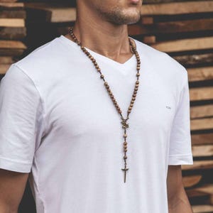May include: A long wooden bead necklace with a bronze-colored cross pendant. The necklace is made of small, round, light brown beads. The pendant is a cross with a detailed design. The necklace is worn over a white V-neck shirt.