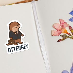 K&ouml;nnte beinhalten: Ein Aufkleber mit einem Cartoon-Otter im Anzug, der eine Aktentasche h&auml;lt, mit dem Wort "OTTERNEY" darunter. Der Aufkleber befindet sich auf einer wei&szlig;en Oberfl&auml;che neben getrockneten Blumen.