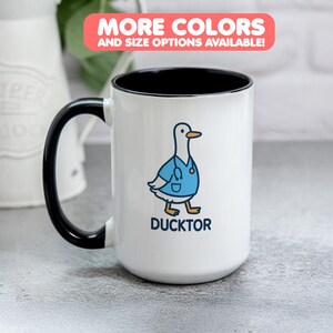 Könnte beinhalten: Weißer Keramikbecher mit schwarzem Griff und Innenrand. Der Becher zeigt eine Cartoon-Ente mit einem blauen Arztkittel und dem Wort "DUCKTOR" darunter. Oben im Bild befindet sich ein rosa Banner mit der Aufschrift "MEHR FARBEN UND GRÖSSENOPTIONEN VERFÜGBAR!"