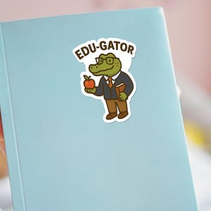 May include: A sticker featuring a cartoon alligator wearing glasses, a suit, and holding an apple and a book. The sticker has the text "EDU-GATOR" in a speech bubble. The sticker is on a light blue notebook.
