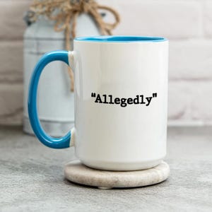 May include: White ceramic mug with a blue interior and handle. The mug features the word "Allegedly" in black text. The mug is sitting on a round, light-colored coaster.