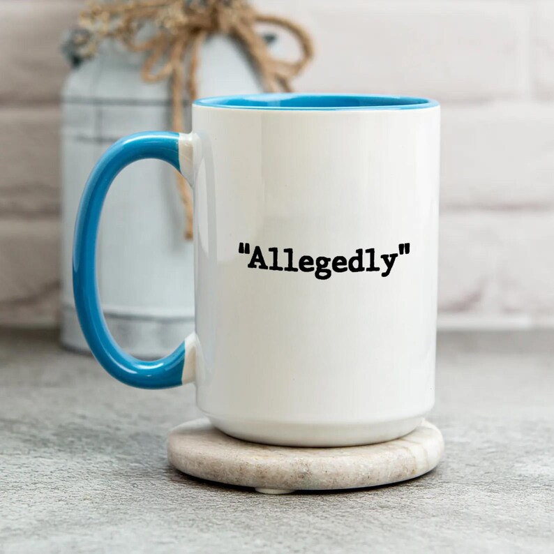 May include: White ceramic mug with a blue interior and handle. The mug features the word "Allegedly" in black text. The mug is sitting on a round, light-colored coaster.