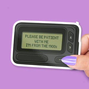 May include: A sticker featuring a black pager with a white border. The pager's screen displays the text "PLEASE BE PATIENT WITH ME I'M FROM THE 1900s". The sticker is held by a hand with a purple painted fingernail against a purple background.