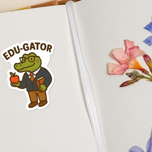 May include: A sticker featuring an alligator character wearing glasses, a suit, and holding an apple and a book. The sticker has the text "EDU-GATOR" above the alligator. The background includes dried flowers and a notebook.