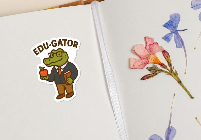 May include: A sticker featuring an alligator character wearing glasses, a suit, and holding an apple and a book. The sticker has the text "EDU-GATOR" above the alligator. The background includes dried flowers and a notebook.