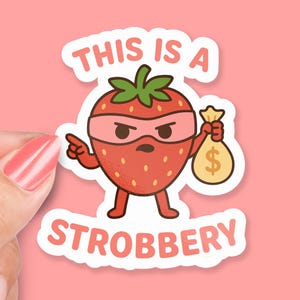 May include: A sticker featuring a cartoon strawberry character wearing a pink mask and holding a money bag with a dollar sign. The text "THIS IS A STROBBERY" is written above and below the strawberry.