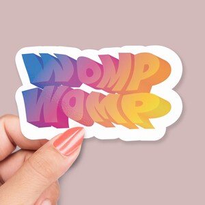 Puede incluir: Una pegatina con el texto "WOMP WOMP" en un degradado arcoíris. El texto está en estilo 3D y está contorneado en blanco.