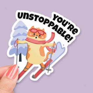 May include: A sticker featuring a cartoon cat wearing ski goggles and a pink scarf, skiing down a snowy mountain. The text "You're unstoppable!" is written in black letters on the sticker.