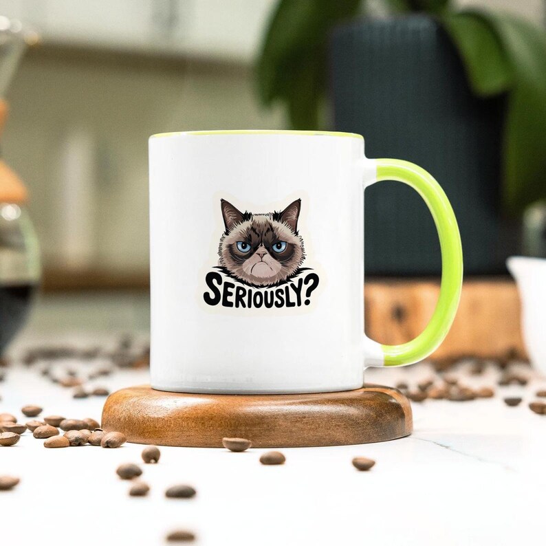 May include: White ceramic mug with a green handle and rim, featuring a grumpy cat graphic and the text "Seriously?". The mug is placed on a wooden coaster, with coffee beans scattered around.