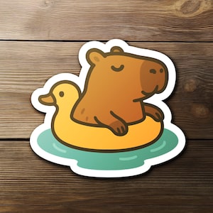 Puede incluir: Una pegatina con un capibara de dibujos animados flotando en un flotador con forma de pato amarillo. El capibara es marrón con una cara marrón claro y brazos pequeños, en agua azul claro.