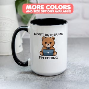 Pode incluir: Caneca de cerâmica branca com alça e interior pretos. A caneca apresenta um urso de desenho animado com uma expressão severa, trabalhando em um laptop. O texto na caneca diz "DON'T BOTHER ME I'M CODING". Uma faixa na parte superior diz "MORE COLORS AND SIZE OPTIONS AVAILABLE!"