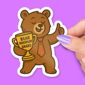 Puede incluir: Una pegatina de oso marrón con un trofeo dorado que dice "BEAR MINIMUM AWARD". El oso lleva una corbata naranja y hace una señal de aprobación. La pegatina está sujeta por una mano con una uña morada.