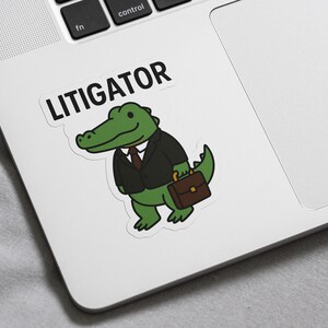 Pode incluir: Um autocolante com um jacar&eacute; de desenho animado de fato, segurando uma maleta. A palavra "LITIGATOR" est&aacute; impressa acima do jacar&eacute; em letras mai&uacute;sculas pretas. O jacar&eacute; &eacute; verde com um fato preto e uma maleta castanha.