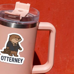 K&ouml;nnte beinhalten: Ein rosa isolierter Becher mit Griff und transparentem Deckel. Auf dem Becher befindet sich ein Aufkleber eines Cartoon-Otters im Anzug mit einer Aktentasche. Der Aufkleber tr&auml;gt das Wort "OTTERNEY" in wei&szlig;er Schrift.