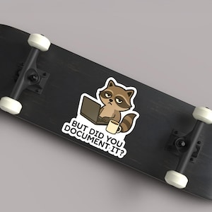 Puede incluir: Una pegatina con un mapache de dibujos animados con un port&aacute;til y una taza de caf&eacute;. El mapache tiene una expresi&oacute;n de mal humor y el texto "BUT DID YOU DOCUMENT IT?" est&aacute; debajo. La pegatina est&aacute; sobre una patineta negra.