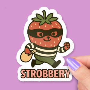 May include: A die-cut sticker featuring a cartoon strawberry character in a black mask and striped shirt, running with a money bag. The word "STROBBERY" is printed in a bold, outlined font below the image. The sticker has a white border and is set against a purple background.