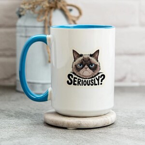 May include: White ceramic mug with a blue interior and handle. The mug features a grumpy cat graphic with the text "SERIOUSLY?" in black. The mug is sitting on a round, beige coaster.