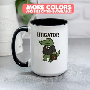 Pode incluir: Caneca de cerâmica branca com alça e interior pretos. A caneca apresenta a palavra "LITIGATOR" acima de um jacaré de desenho animado usando um terno e segurando uma pasta. O texto na parte superior diz "MORE COLORS AND SIZE OPTIONS AVAILABLE!"