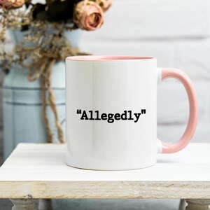 May include: White ceramic mug with a pink interior and handle. The mug features the word "Allegedly" in black, centered on the front. The mug is set on a white surface, with a blurred background of flowers and a vase.