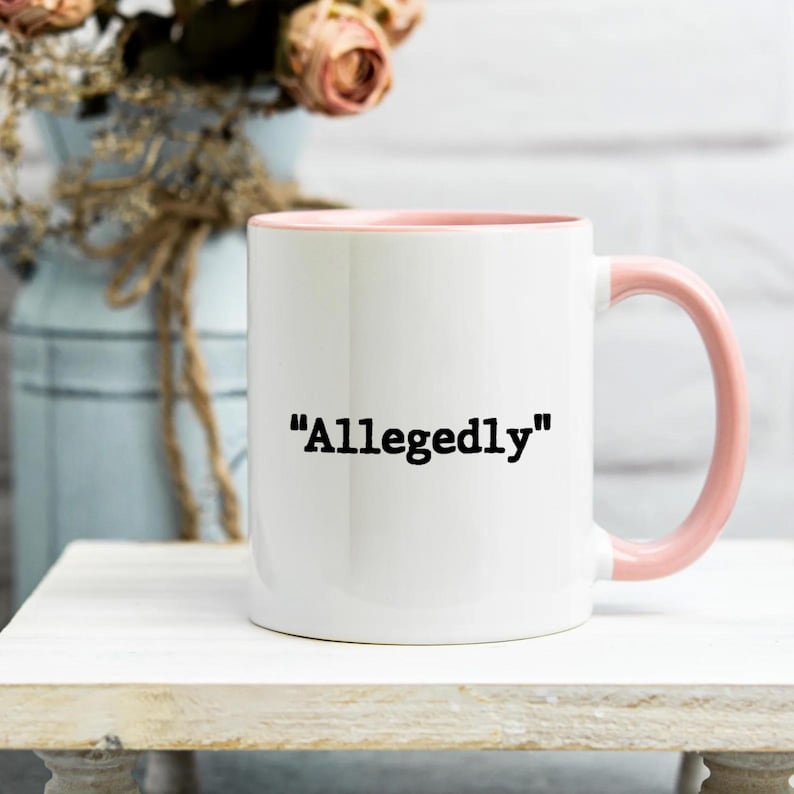 May include: White ceramic mug with a pink interior and handle. The mug features the word "Allegedly" in black, centered on the front. The mug is set on a white surface, with a blurred background of flowers and a vase.