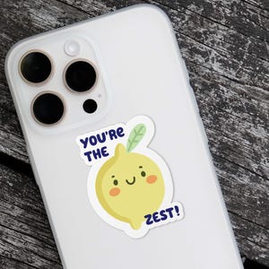 May include: A white phone case with a yellow lemon sticker that says "You're the Zest!" The lemon has a smiling face and a green leaf.