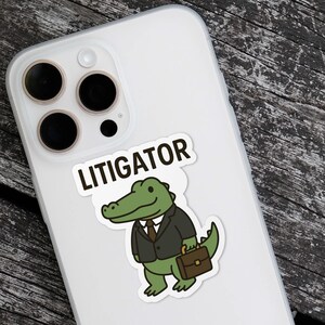 Pode incluir: Um autocolante branco com a palavra "LITIGATOR" acima de um jacar&eacute; de desenho animado usando um terno e segurando uma pasta. O jacar&eacute; &eacute; verde com uma gravata marrom e um terno preto. O autocolante est&aacute; em uma capa de telefone branca.