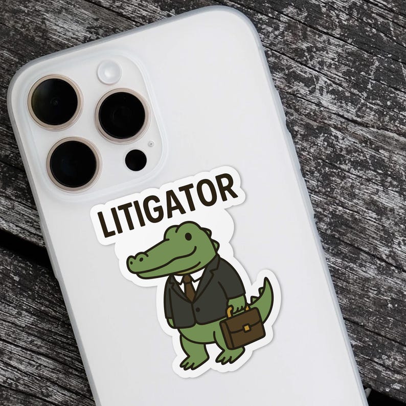 Pode incluir: Um autocolante branco com a palavra "LITIGATOR" acima de um jacar&eacute; de desenho animado usando um terno e segurando uma pasta. O jacar&eacute; &eacute; verde com uma gravata marrom e um terno preto. O autocolante est&aacute; em uma capa de telefone branca.